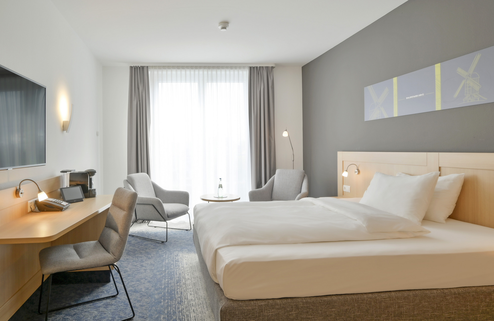 Stilvolles Hotelzimmer mit Bett, Schreibtisch, Stuhl, Fernseher, grauen Sesseln und großem Fenster
