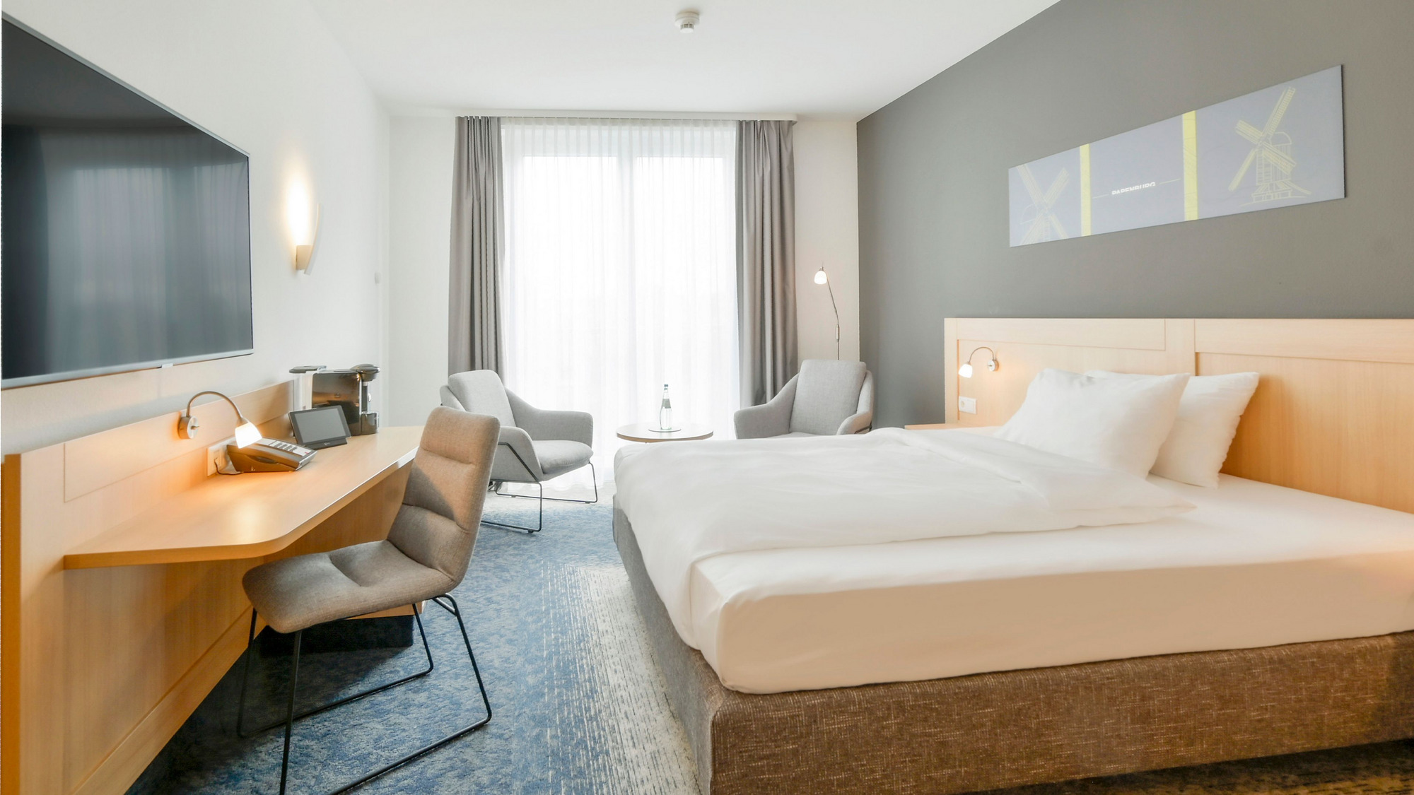 Stilvolles Hotelzimmer mit einem komfortablen Bett, einem Schreibtisch, einem Stuhl, einem Fernseher, zwei grauen Sesseln mit Beistelltisch und großem Fenster