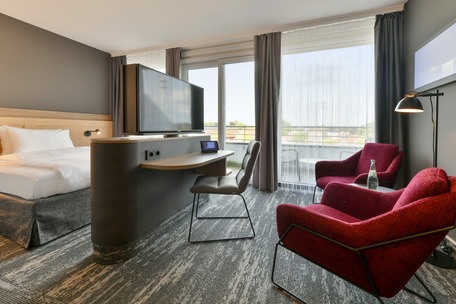 Stilvolles Hotelzimmer mit Balkon, Regal mit Fernseher, Schreibtisch, Bett und Sitzecke mit roten Sesseln