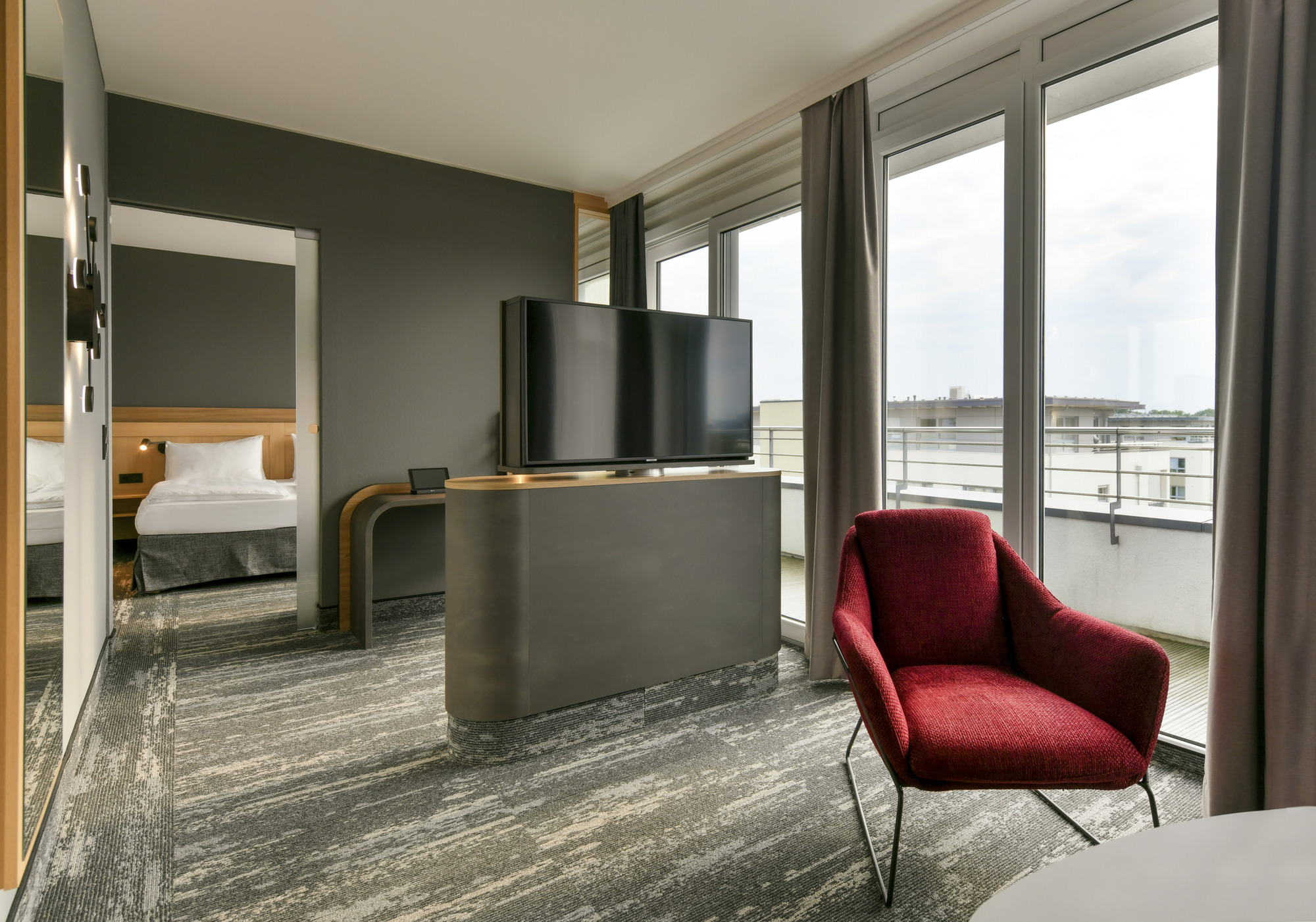 Modernes Hotelzimmer mit einem Fernseher, einem roten Sessel mit Beistelltisch und einem weitläufigen Balkon, im Hintergrund ist ein separares Schlafzimmer zu erkennen